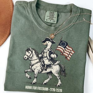 May include: Green Comfort Colors t-shirt featuring a cartoon duck riding a horse and holding a flag. The duck is dressed in colonial attire. The shirt has the text "HONK FOR FREEDOM - 1776-2026" at the bottom. A gold necklace is draped across the shirt.