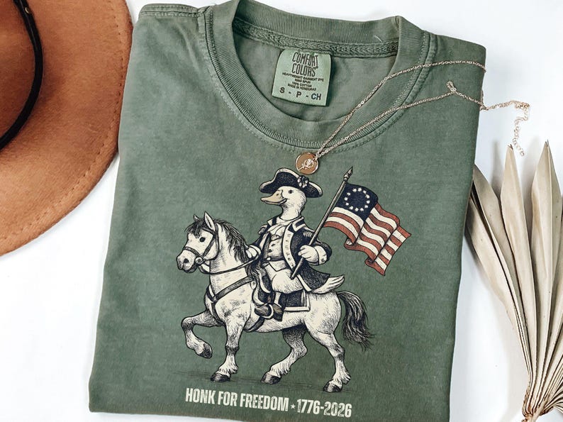 May include: Green Comfort Colors t-shirt featuring a cartoon duck riding a horse and holding a flag. The duck is dressed in colonial attire. The shirt has the text "HONK FOR FREEDOM - 1776-2026" at the bottom. A gold necklace is draped across the shirt.