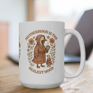 Puede incluir: Taza de caf&eacute; de cer&aacute;mica blanca con una ilustraci&oacute;n marr&oacute;n y blanca de un ganso con una bata y sosteniendo una taza de caf&eacute;. El texto "MOTHERHOOD IS THE REALEST HOOD" est&aacute; impreso en la taza.