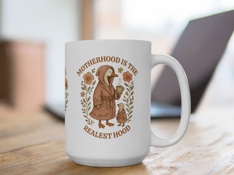 Puede incluir: Taza de caf&eacute; de cer&aacute;mica blanca con una ilustraci&oacute;n marr&oacute;n y blanca de un ganso con una bata y sosteniendo una taza de caf&eacute;. El texto "MOTHERHOOD IS THE REALEST HOOD" est&aacute; impreso en la taza.