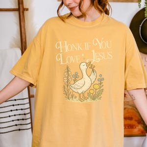 Puede incluir: Camiseta de color amarillo mostaza con la frase "Honk If You Love Jesus". El dise&ntilde;o incluye un pato sosteniendo una cruz, rodeado de flores y hierba. La camiseta es de estilo casual y c&oacute;modo.