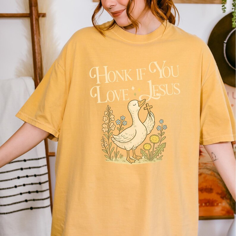 Puede incluir: Camiseta de color amarillo mostaza con la frase "Honk If You Love Jesus". El dise&ntilde;o incluye un pato sosteniendo una cruz, rodeado de flores y hierba. La camiseta es de estilo casual y c&oacute;modo.