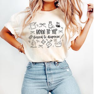 May include: Cream-colored t-shirt with the text "BORN TO YAP forced to dispense" in black, surrounded by small medical-themed illustrations. The shirt is paired with blue jeans.