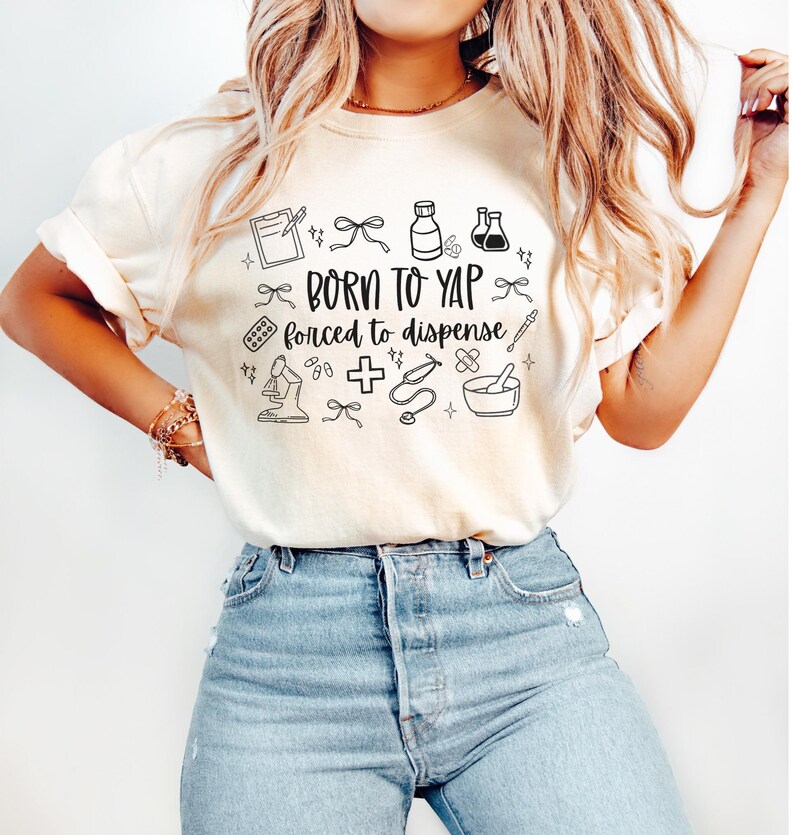 May include: Cream-colored t-shirt with the text "BORN TO YAP forced to dispense" in black, surrounded by small medical-themed illustrations. The shirt is paired with blue jeans.