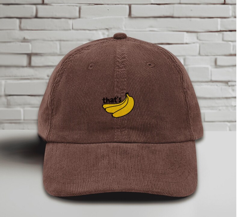 Brown baseball cap with a curved brim featuring an embroidered yellow banana design and the word “that’s” stitched in black above it. The hat is displayed upright against a white brick wall background, showcasing its soft, unstructured style and casual look.