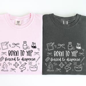 May include: Two t-shirts, one pink and one charcoal gray, are displayed. Each shirt features the phrase "BORN TO YAP forced to dispense" in a whimsical font, surrounded by medical-themed illustrations in white.