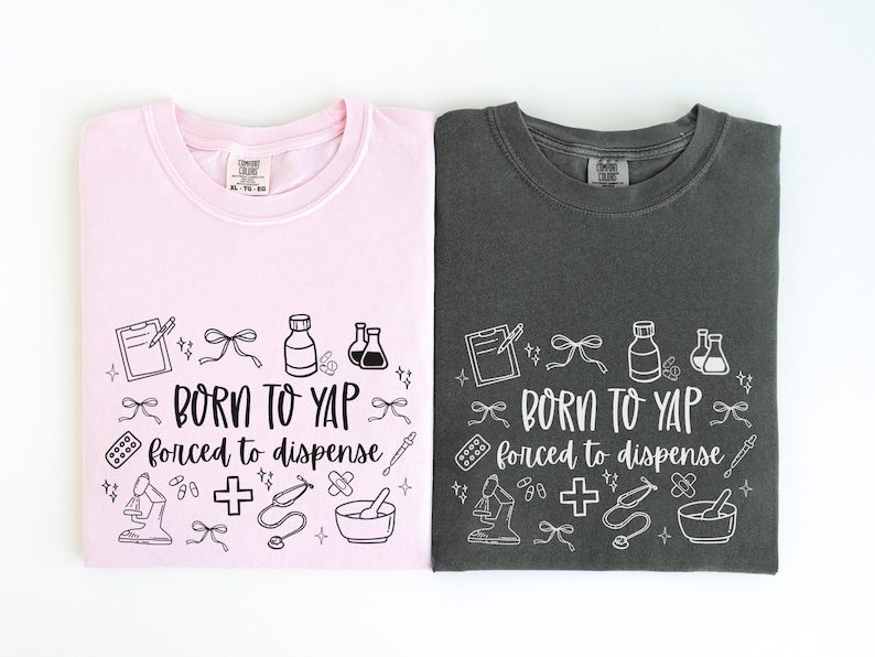 May include: Two t-shirts, one pink and one charcoal gray, are displayed. Each shirt features the phrase "BORN TO YAP forced to dispense" in a whimsical font, surrounded by medical-themed illustrations in white.