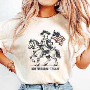 May include: Cream-colored t-shirt featuring a cartoon duck riding a horse, dressed in colonial attire, and holding a flag. The flag has a circle of stars and stripes. The text "HONK FOR FREEDOM - 1776-2026" is printed below the image.