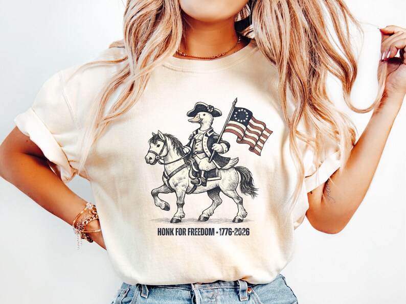 May include: Cream-colored t-shirt featuring a cartoon duck riding a horse, dressed in colonial attire, and holding a flag. The flag has a circle of stars and stripes. The text "HONK FOR FREEDOM - 1776-2026" is printed below the image.