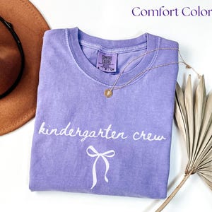 Folded lavender Comfort Colors t-shirt displayed on a white surface with a neutral hat and dried palm leaves. The shirt features the phrase “kindergarten crew” in white cursive with a small bow illustration underneath. Styled with a gold pendant necklace draped over the shirt for a soft, feminine, teacher-themed flat lay perfect for back-to-school season.