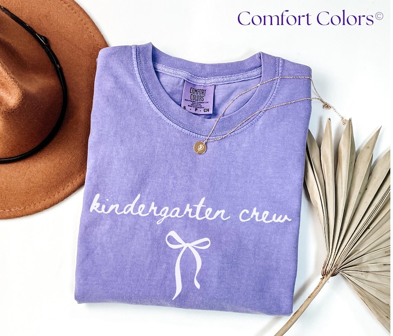 Folded lavender Comfort Colors t-shirt displayed on a white surface with a neutral hat and dried palm leaves. The shirt features the phrase “kindergarten crew” in white cursive with a small bow illustration underneath. Styled with a gold pendant necklace draped over the shirt for a soft, feminine, teacher-themed flat lay perfect for back-to-school season.