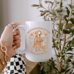 Puede incluir: Taza de cer&aacute;mica blanca con una ilustraci&oacute;n de dibujos animados de un pato con una bata y sosteniendo una taza. El texto "Motherhood is the Realest Hood" est&aacute; impreso en la taza.
