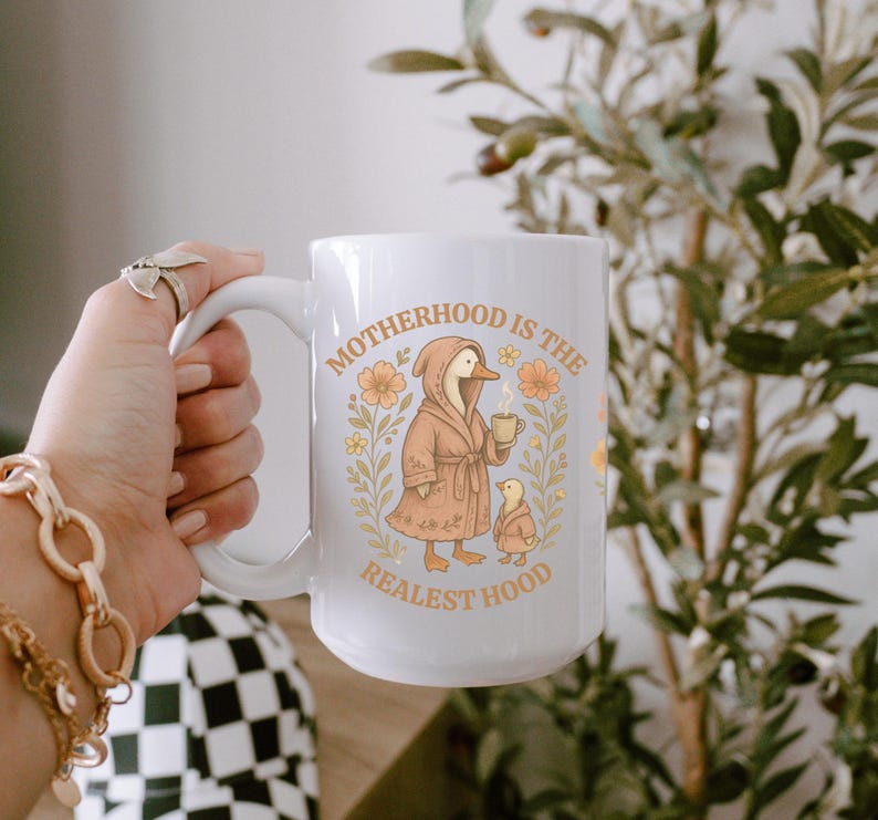 Puede incluir: Taza de cer&aacute;mica blanca con una ilustraci&oacute;n de dibujos animados de un pato con una bata y sosteniendo una taza. El texto "Motherhood is the Realest Hood" est&aacute; impreso en la taza.