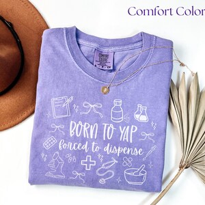 May include: A lilac Comfort Colors t-shirt with the text "BORN TO YAP forced to dispense" and medical-themed graphics in white. A gold necklace and a brown hat are partially visible in the image.