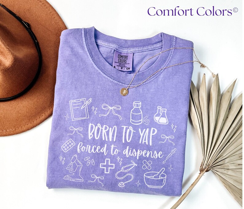 May include: A lilac Comfort Colors t-shirt with the text "BORN TO YAP forced to dispense" and medical-themed graphics in white. A gold necklace and a brown hat are partially visible in the image.