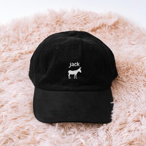 May include: Black corduroy baseball cap with a curved brim. The cap features the name "jack" embroidered in white above a white donkey silhouette. The hat is displayed on a fluffy, light pink surface.