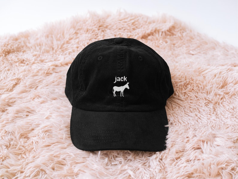 May include: Black corduroy baseball cap with a curved brim. The cap features the name "jack" embroidered in white above a white donkey silhouette. The hat is displayed on a fluffy, light pink surface.