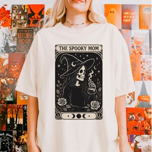 May include: Cream-colored t-shirt featuring a black and white graphic of a skeleton witch holding a potion bottle, with the text "THE SPOOKY MOM" above the image. The design includes roses, moons, and stars, creating a Halloween theme.