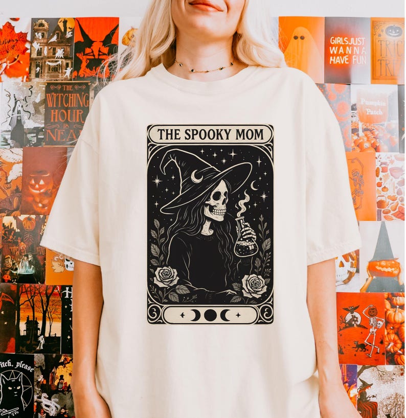 May include: Cream-colored t-shirt featuring a black and white graphic of a skeleton witch holding a potion bottle, with the text "THE SPOOKY MOM" above the image. The design includes roses, moons, and stars, creating a Halloween theme.