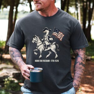 May include: Dark gray t-shirt featuring a cartoon duck riding a horse, dressed in colonial attire and holding a flag. The flag has 13 stars. The text "HONK FOR FREEDOM - 1776-2026" is printed below the image.