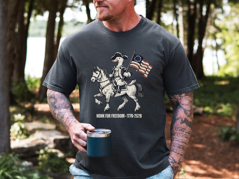 May include: Dark gray t-shirt featuring a cartoon duck riding a horse, dressed in colonial attire and holding a flag. The flag has 13 stars. The text "HONK FOR FREEDOM - 1776-2026" is printed below the image.