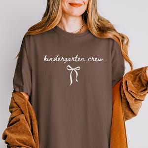 May include: A brown t-shirt with the words "kindergarten crew" in a cursive font and a white bow graphic. The shirt is part of a clothing collection.