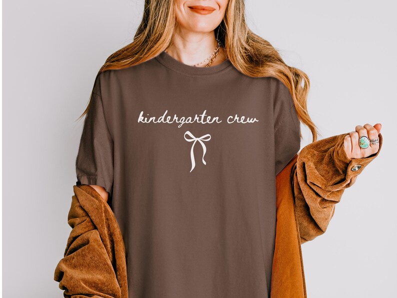 May include: A brown t-shirt with the words "kindergarten crew" in a cursive font and a white bow graphic. The shirt is part of a clothing collection.