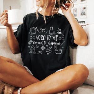 May include: Black t-shirt with white text and medical-themed illustrations. The shirt reads "Born to Yap, forced to dispense." The design includes a clipboard, pills, a microscope, and a mortar and pestle.