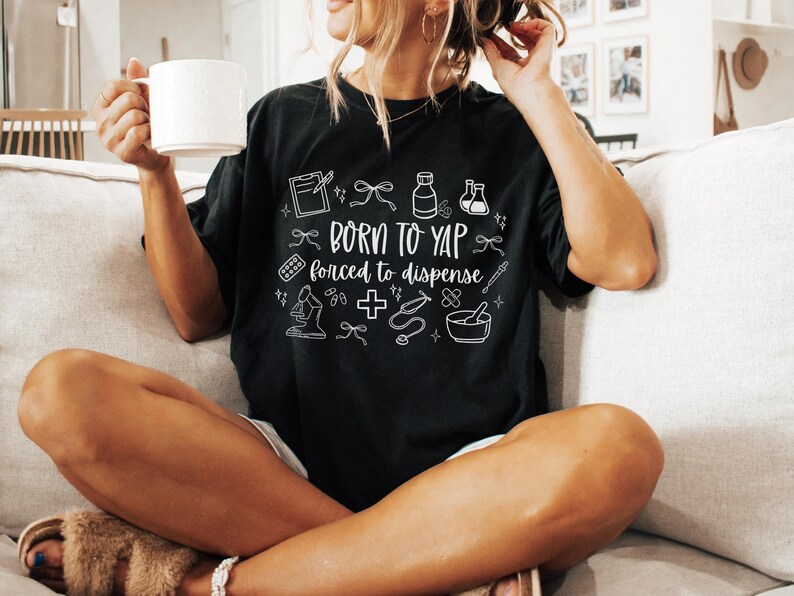 May include: Black t-shirt with white text and medical-themed illustrations. The shirt reads "Born to Yap, forced to dispense." The design includes a clipboard, pills, a microscope, and a mortar and pestle.