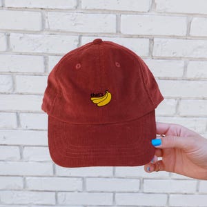 Puede incluir: Una gorra de b&eacute;isbol de pana roja con el texto "That's Bananas" bordado en amarillo en la parte delantera. La gorra tiene una visera curvada.