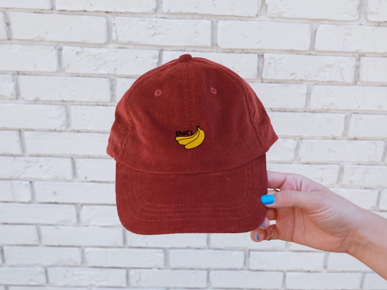 Puede incluir: Una gorra de b&eacute;isbol de pana roja con el texto "That's Bananas" bordado en amarillo en la parte delantera. La gorra tiene una visera curvada.
