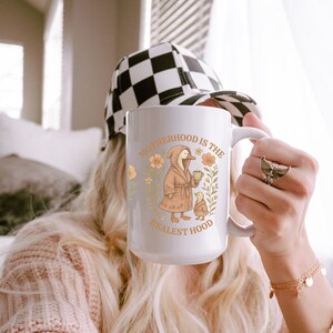 Puede incluir: Taza de cer&aacute;mica blanca con un dise&ntilde;o floral y un pato de dibujos animados con una bata y sosteniendo una taza. El texto en la taza dice "Motherhood is the Realest Hood".