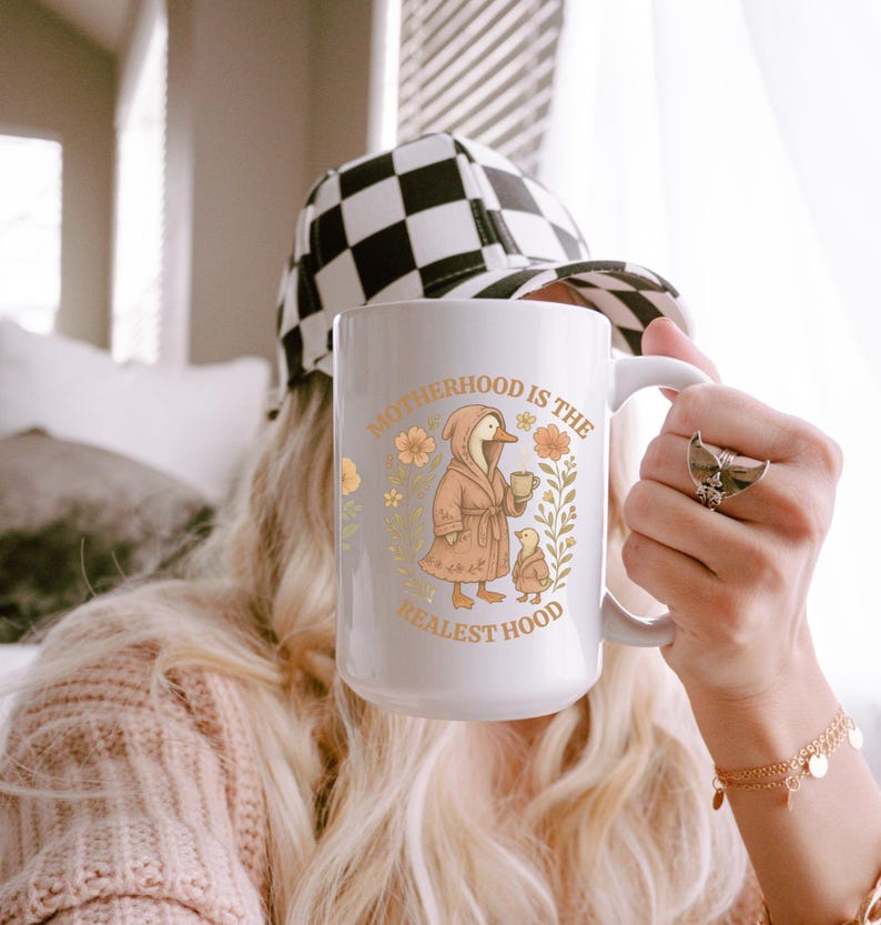 Puede incluir: Taza de cer&aacute;mica blanca con un dise&ntilde;o floral y un pato de dibujos animados con una bata y sosteniendo una taza. El texto en la taza dice "Motherhood is the Realest Hood".
