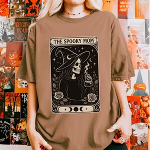 May include: A brown t-shirt featuring a black and white graphic of a skeleton witch holding a potion bottle, with the text "THE SPOOKY MOM" in a decorative frame. The design includes stars, roses, and moon phases, perfect for Halloween.