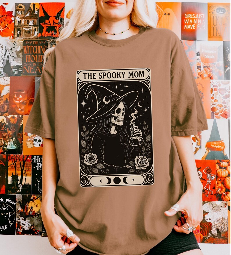 May include: A brown t-shirt featuring a black and white graphic of a skeleton witch holding a potion bottle, with the text "THE SPOOKY MOM" in a decorative frame. The design includes stars, roses, and moon phases, perfect for Halloween.