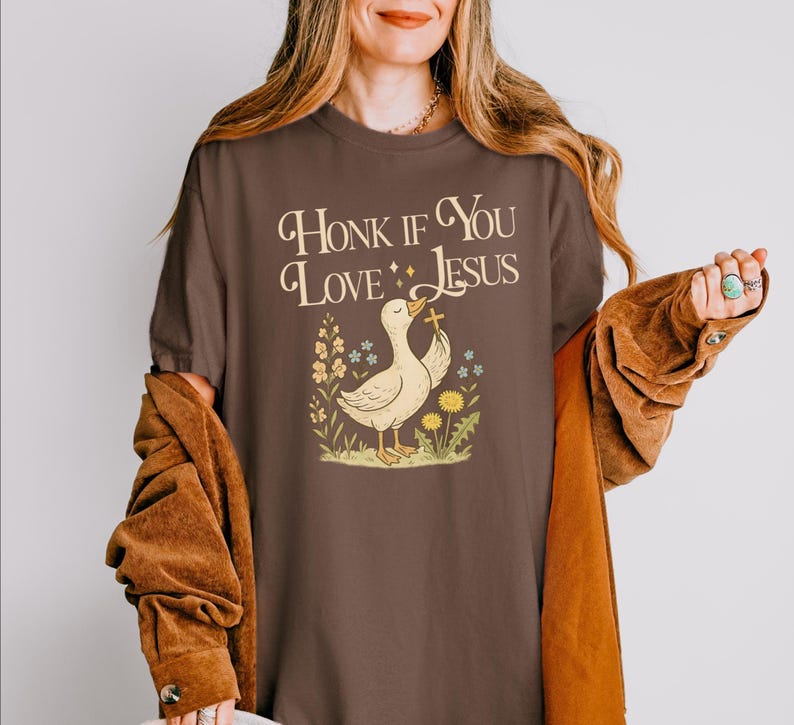Puede incluir: Camiseta marr&oacute;n con el texto "Honk If You Love Jesus" sobre un pato de dibujos animados sosteniendo una cruz. El pato est&aacute; rodeado de flores. La camiseta es de color marr&oacute;n liso.
