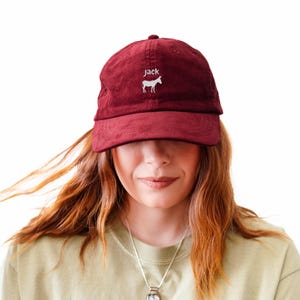 May include: A burgundy corduroy baseball cap with the name "jack" embroidered in white above a white horse graphic. The cap has a curved brim and a fabric strap closure. The cap is worn by a person.