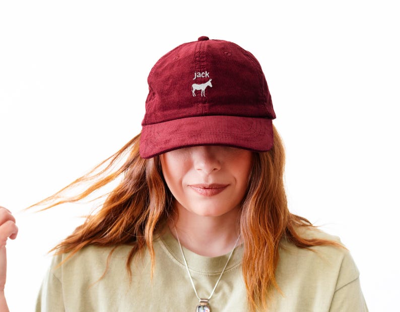 May include: A burgundy corduroy baseball cap with the name "jack" embroidered in white above a white horse graphic. The cap has a curved brim and a fabric strap closure. The cap is worn by a person.