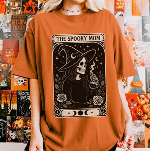 May include: Orange t-shirt featuring a black and white graphic of a skeleton wearing a witch hat, holding a flask, and surrounded by roses. The text "THE SPOOKY MOM" is displayed above the graphic. The shirt is a Halloween-themed item.
