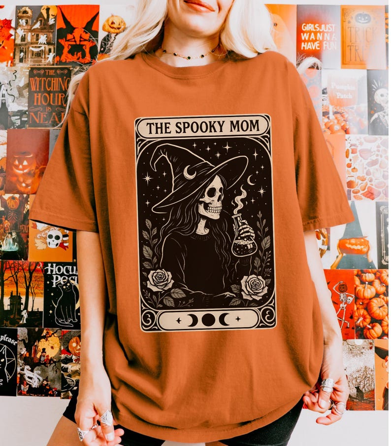 May include: Orange t-shirt featuring a black and white graphic of a skeleton wearing a witch hat, holding a flask, and surrounded by roses. The text "THE SPOOKY MOM" is displayed above the graphic. The shirt is a Halloween-themed item.