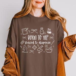 May include: A brown t-shirt with the text "BORN TO YAP forced to dispense" in white. The shirt features white line drawings of medical and pharmacy-related items, such as a clipboard, pills, and a mortar and pestle.