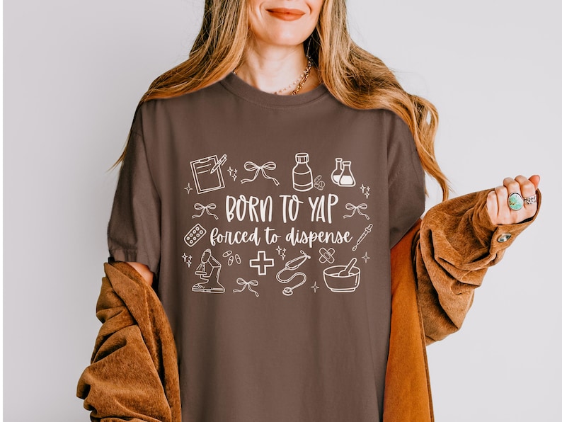 May include: A brown t-shirt with the text "BORN TO YAP forced to dispense" in white. The shirt features white line drawings of medical and pharmacy-related items, such as a clipboard, pills, and a mortar and pestle.