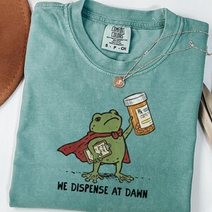 May include: A sage green t-shirt featuring a graphic of a green frog wearing a red cape, holding a clipboard and a prescription pill bottle. The text below the frog reads, "WE DISPENSE AT DAWN." The shirt has a Comfort Colors brand label.