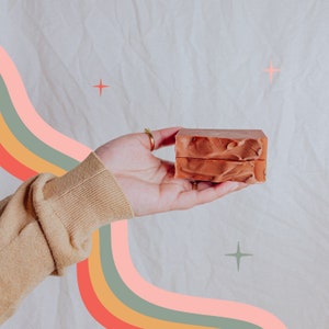 May include: Two rectangular bars of reddish-brown soap are held in a hand. The soap has a textured surface. The background is a light beige with a rainbow-coloured arc and starbursts.