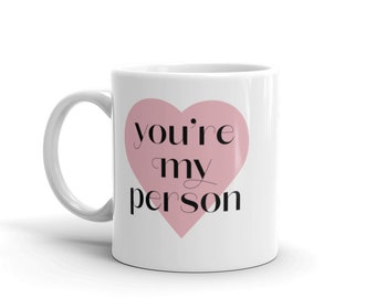 Può includere: Tazza in ceramica bianca con manico curvo. La tazza presenta un cuore rosa con le parole "you're my person" in corsivo nero. La tazza è progettata per le bevande.