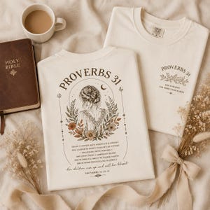 May include: An off-white crewneck t-shirt featuring a back graphic of a woman surrounded by flowers and the text Proverbs 31, along with verses from the Bible. A smaller matching graphic is printed on the front chest area of the folded shirt.
