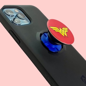 Könnte beinhalten: Schwarze Smartphone-Hülle mit einem roten und gelben Wonder Woman Logo-Pop-Socket. Der Pop-Socket hat eine blaue Basis. Die Handyhülle befindet sich auf einem rosa-grünen Hintergrund.