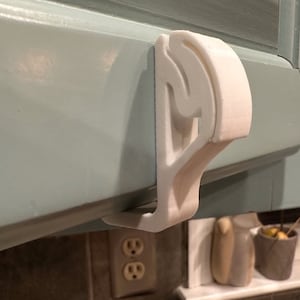 May include: White plastic hook designed to be mounted under a cabinet. The hook is shaped like a letter 'J' and has a curved top.