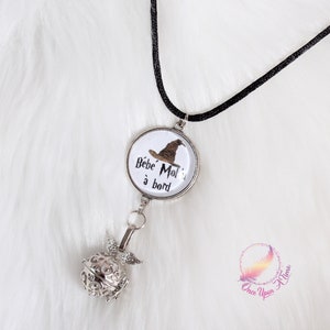 Puede incluir: Collar plateado con cordón negro. El colgante presenta un fondo blanco con un gráfico de sombrero marrón y el texto "Bébé Moi à bord". Una esfera decorativa calada cuelga debajo del colgante.