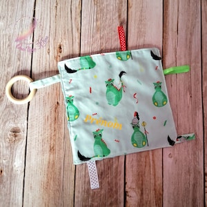 Op de afbeelding: Een lichtblauw babyzintuiglijk speelgoed met een houten ring, linten en een vierkant van stof met een groen drakenpatroon. Het vierkant van stof is geborduurd met het woord "Prénom".